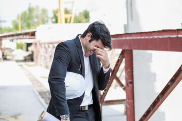 Portrait of Hispanic male engineer in suit looking stressed and frustrated at construction site, holding safety helmet and rolled blueprint under arm in natural daylight.
