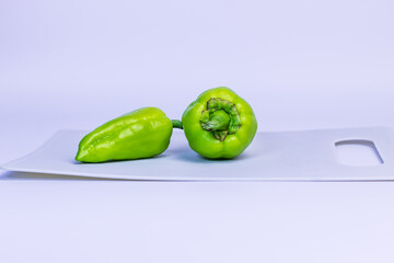 Two green bell peppers lie on a light gray cutting board. The peppers are fresh, one is positioned vertically and the other is lying on its side, highlighting their bright color.