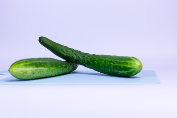 Two fresh, long green cucumbers are arranged in a cross shape on a light blue cutting board. The cutting board has a hole for a handle on a white background. The cucumbers have a bumpy texture.