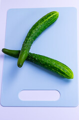 Two fresh, long green cucumbers are arranged in a cross shape on a light blue cutting board. The cutting board has a hole for a handle on a white background. The cucumbers have a bumpy texture.