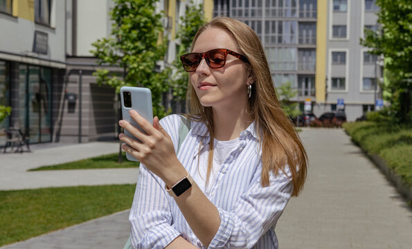 Young woman with long blonde hair is standing outside, smiling, looking at her smartphone. She is wearing casual clothes and has a smartwatch. Green trees and city buildings are visible in the back. - Powered by Adobe