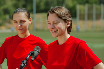 Smiling women football players giving interview on field in red jerseys. Concept of positivity in female athletics, smiling footballers during interview, promoting youth confidence.