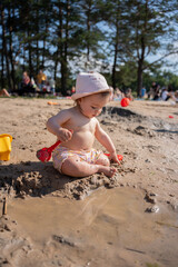 a young child playing joyfully on a sandy beach, using a red shovel to explore and create in the mud, surrounded by the natural beauty of trees and a sunny day's warmth, youth, environment, scene