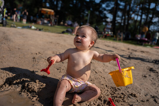 joyful baby with bright yellow bucket and red plastic spade plays in a sandy outdoor area surrounded by blurred green park environment, basking in warm, cheerful sunlight, interaction, joy, light