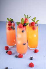 vibrant refreshing beverages in tall glasses garnished with fresh raspberries, blueberries, and mint leaves set against a soft blue background evoking a zesty summertime vibe, minimalist, stylish