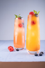 vibrant fruit-infused summer cocktails in tall glasses with ice, garnished with fresh raspberries, blueberries, and mint leaves on a cool, light blue background surface under soft lighting