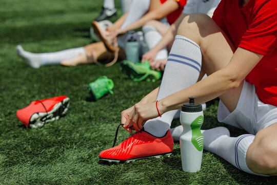 Female soccer athletes getting ready for game on sunlit field. Concept of self-care during practice, close-up on hydration and shoes, healthy routines in athletics.