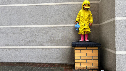 Child in bright yellow raincoat and pink boots standing on a low brick platform against a grey wall, holding a blue watering can. Outdoor playful moment in rainy weather.