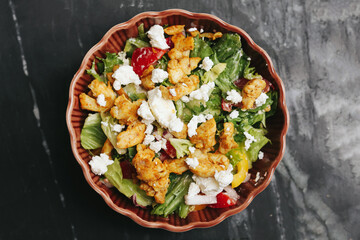Fresh salad in ceramic bowl. Healthy lunch with lettuce, fried chicken, feta cheese and cherry tomatoes. Colorful protein-rich meal on dark marble background. Tasty and nutritious food idea.