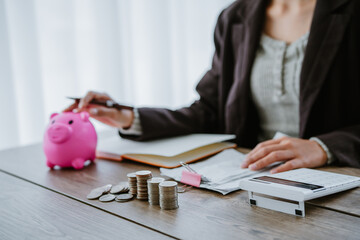 Woman placing coins into a piggy bank, with stacks of coins, calculator, and notebook nearby, symbolizing saving and financial planning