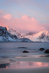 Colorful winter sunrise at Haukland Beach, Lofoten Islands, Norway. Snowy mountain and pink clouds reflect in the calm sea and wet sand, creating a peaceful Arctic landscape.