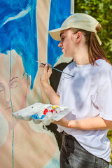 Young female artist with fair hair wearing cap paints large canvas outside. Blue dominates artwork....
