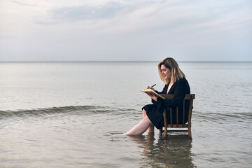 Young woman sits on chair in shallow water at sunset, writing in notebook. Serene ocean setting...