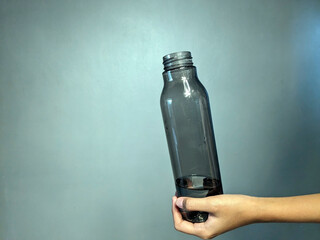 A person's hand holding a transparent dark gray plastic water bottle tilted with a small amount of...