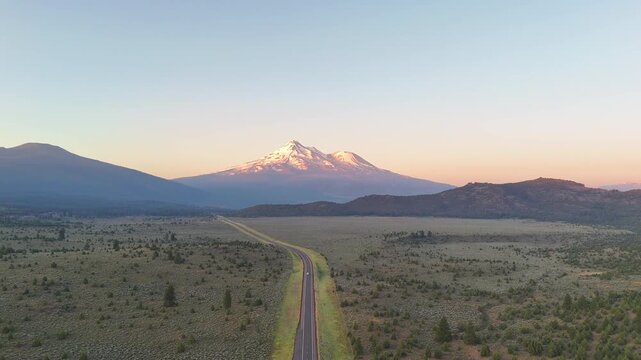 Aerial view of highway 97 leading to snow-capped Mount Shasta, a landmark rising against an orange and yellow morning sky, surrounded by arid vegetation, Mount Shasta, California, United States.