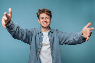 A young man with a friendly smile extends his arms for a hug against a blue background.