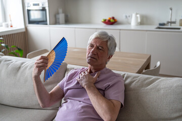 Elderly man using hand fan to cool down in sweltering apartment. Heat exhaustion among seniors,...