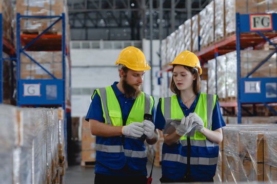 Two warehouse workers wearing safety vests and helmets scanning product barcodes in storage facility, showing modern inventory management and teamwork.