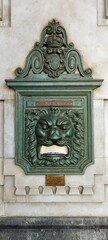 A decorative lion head with a metal door is mounted on a wall. The lion head is made of metal and has a green color. The door is also made of metal and has a unique design