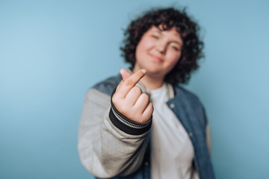 A person with curly hair makes a finger heart gesture against a blue background.