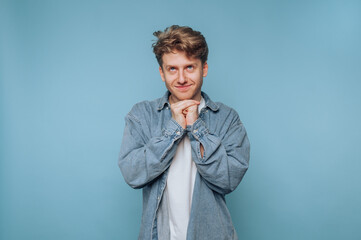 Young man in denim shirt smiling with hands clasped under chin against blue background.