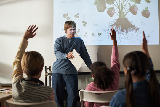 Teenage Caucasian boy standing in front of classroom holding paper, smiling and engaging with diverse group of children raising hands, botanical diagram projected on wall