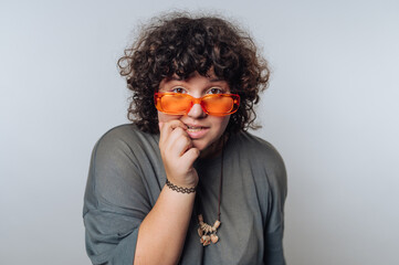 A person with curly hair wearing orange sunglasses and a casual gray shirt, posing playfully...
