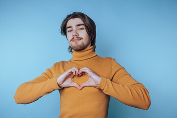 Young man in mustard turtleneck making heart shape with hands against blue background.