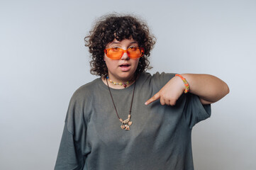 A person with curly hair wearing orange sunglasses and pointing at a necklace.