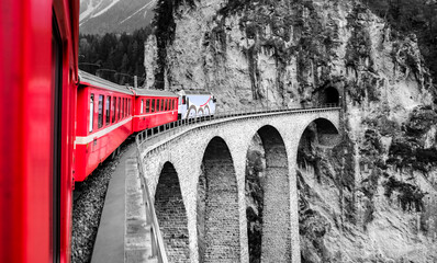 View from the window of a train crossing the Landwasser Viaduct (Switzerland) just before the tunnel. The narrow-gauge railway line is a World Heritage Site. Black-and-white landscape with a red train