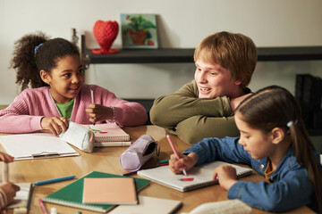 Caucasian teenage boy interacting with Black girl and Caucasian girl while sitting at table, all drawing or writing in notebooks, children and teenager engaging in creative activity together