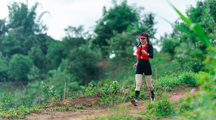 A woman wearing athletic gear and a cap hikes on a dirt trail surrounded by lush greenery and trees under a bright sky.