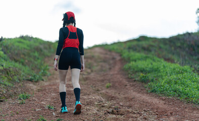 A person wearing athletic gear and a hydration pack walks up a dirt trail surrounded by greenery...