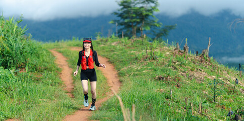 A person trails running on a dirt path surrounded by green vegetation and mountains under a cloudy sky.