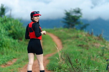 A woman in sporty attire smiles while hiking along a dirt trail in a lush green landscape with...