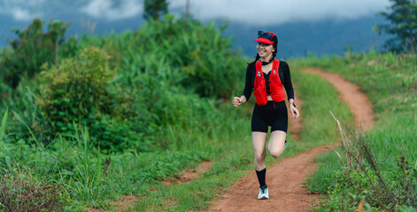 A woman in athletic gear runs happily on a dirt trail surrounded by lush greenery and hills in the...