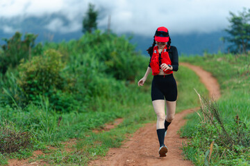 Woman trail running on a dirt path surrounded by greenery under a cloudy sky, wearing athletic gear...