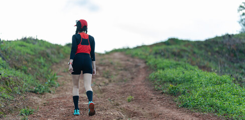 A person wearing athletic gear hikes up a dirt trail surrounded by green hills under a bright sky.
