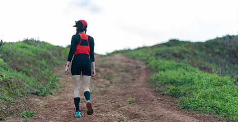 A person in athletic wear and a red cap walks uphill on a dirt path surrounded by sparse greenery under a cloudy sky.
