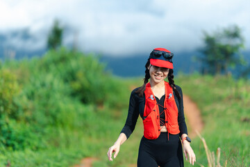 A woman wearing a red cap and vest walks happily along a dirt trail surrounded by lush greenery under a cloudy sky.