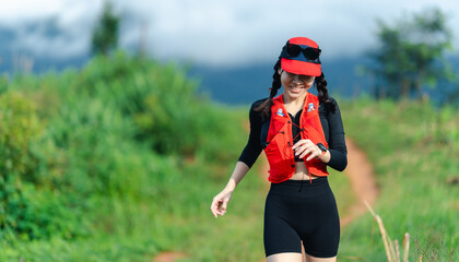 A smiling woman in athletic wear jogs along a green trail under a cloudy sky, wearing a red cap and life vest, enjoying an outdoor run.