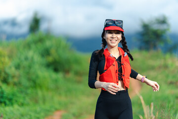 A smiling woman wearing a red vest and cap runs outdoors on a trail surrounded by greenery under a cloudy sky.