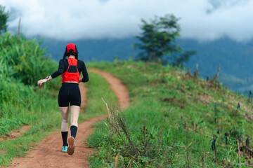 A person in athletic gear runs uphill on a dirt trail surrounded by greenery and misty mountains.