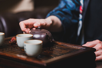 Close-up of a traditional tea ceremony showing a hand pouring tea from a brown teapot into small white cups on a wooden tray, capturing cultural elegance and ritual detail.