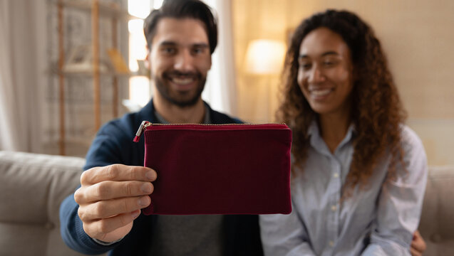 A smiling couple is holding up a small red pouch, possibly a gift, in a cozy living room setting