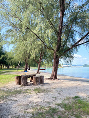 A peaceful beach scene with a wooden table and seats under tall trees, overlooking calm blue waters and distant mountains, ideal for a tranquil escape in Malaysia.