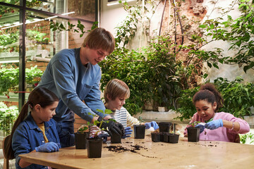 Caucasian young adult man assisting two girls and one Caucasian boy with planting seedlings in...