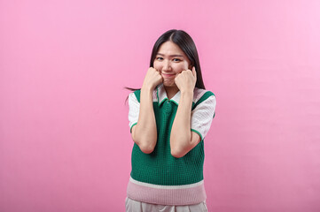 Asian woman with a playful expression poses with clenched fists under her chin, wearing a green...