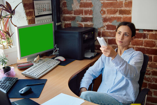 Woman using air conditioner remote in modern office with brick wall. Green screen computer monitor. Concept of office cooling problems, remote work comfort, air conditioning service request.