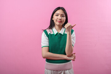 Asian woman in green knit vest pointing her thumb over her shoulder with a doubtful smirk, posing against a solid pink background
